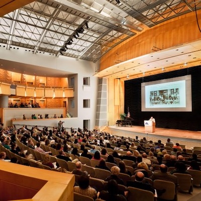 Large audience seated in an amphitheater listening to a speaker at a podium with a presentation projected on screen.