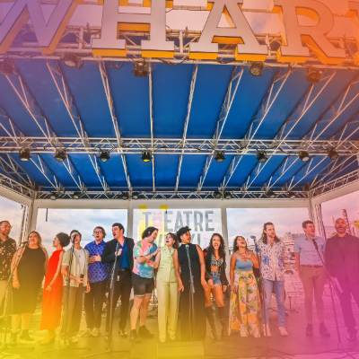 Group of performers standing and singing on stage under a “WHARF” sign during Theatre Week.