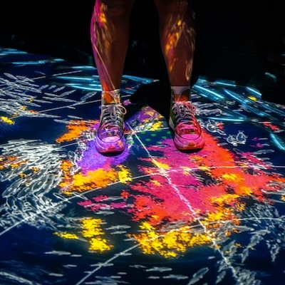 A pair of legs stands on an interactive art installation at ARTECHOUSE in Washington, DC, with vibrant neon patterns and colors projected onto the floor.