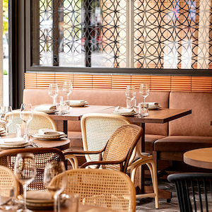 Sunlit restaurant interior with cane-back chairs and neatly set wooden tables beside a cushioned booth.
