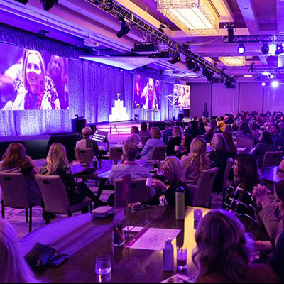A large audience watches a speaker on stage at a professional indoor conference with purple lighting and multiple video screens displaying attendees.