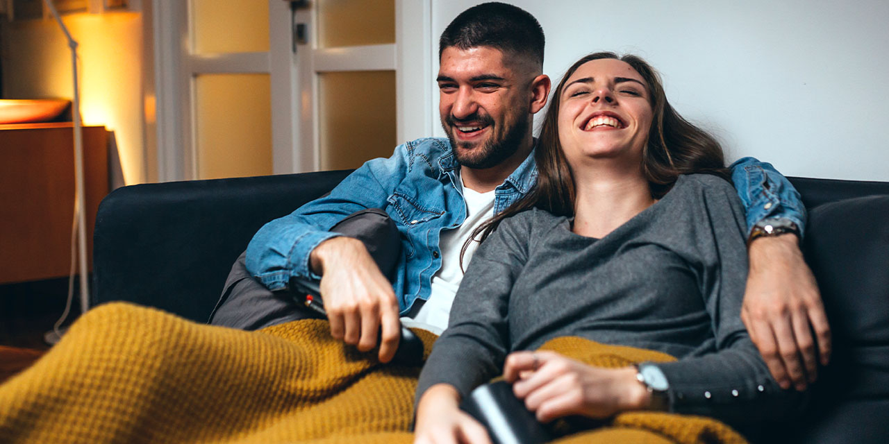 A couple relaxes on a couch under a cozy blanket, laughing together while holding a TV remote.