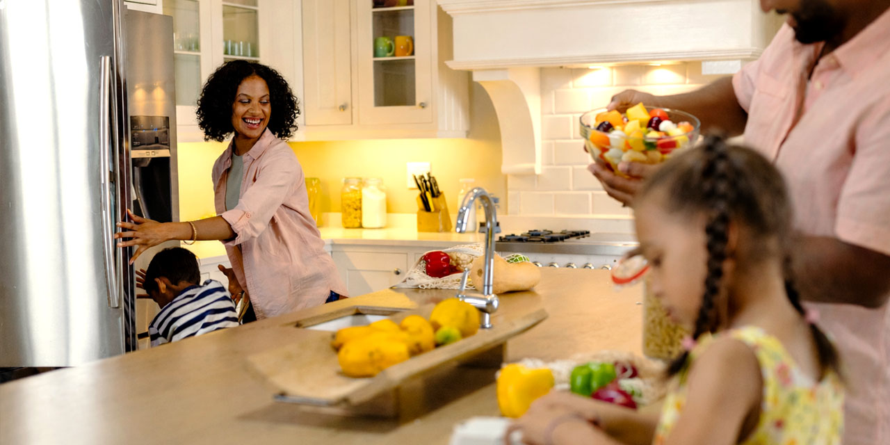 Smiling mom opens the refrigerator while dad holds a bowl of fresh fruit and their children play at the kitchen island in a bright, modern home