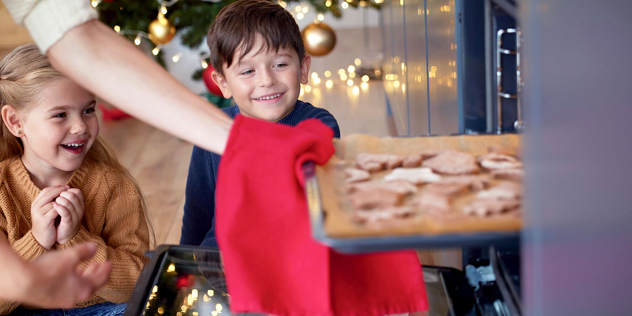 Two excited children sit by an open oven watching a tray of holiday cookies being pulled out