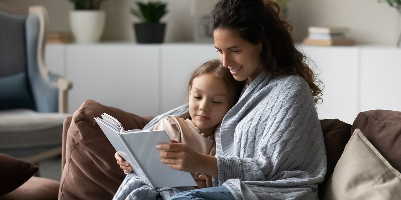 Mother and young daughter cuddle under a blanket on the couch reading a book together in a cozy living room