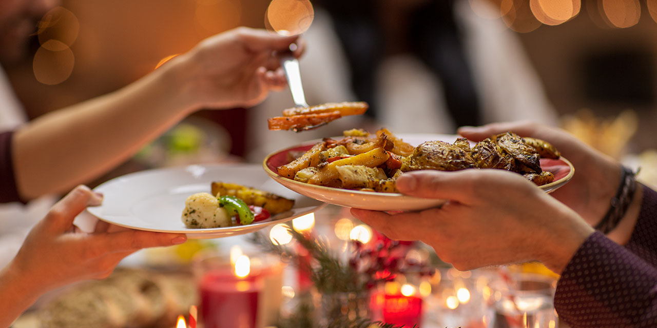 Hands serve roasted holiday vegetables from a platter at a festive candlelit dinner table