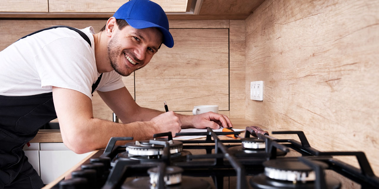 Smiling technician in black overalls and a blue cap writes notes beside a gas cooktop in a modern kitchen