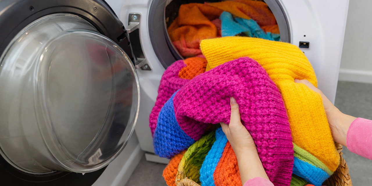 Hands load bright, thick knit sweaters from a basket into an open front-loading washer