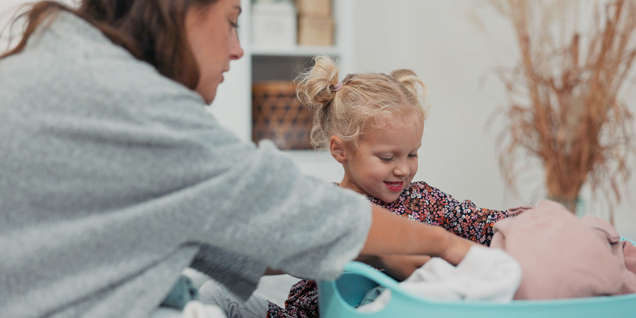 A woman helps a smiling young girl fold clothes into a laundry basket at home