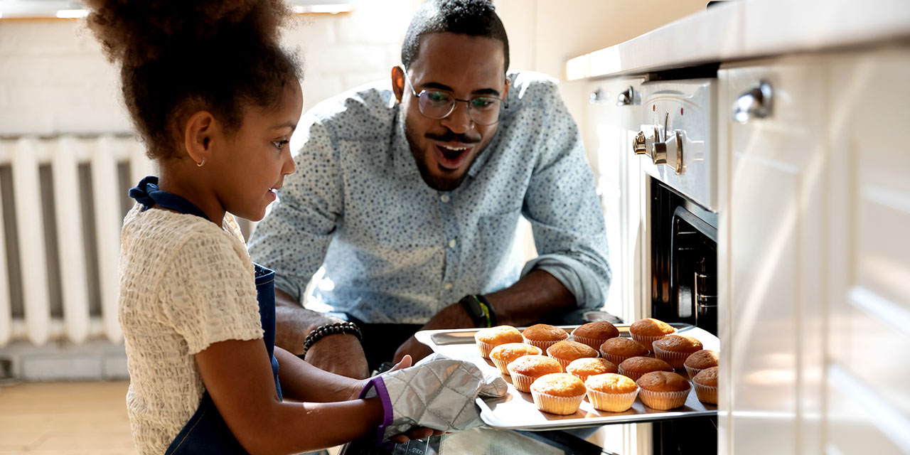 An adult and child pull a tray of freshly baked muffins from the oven in a bright kitchen