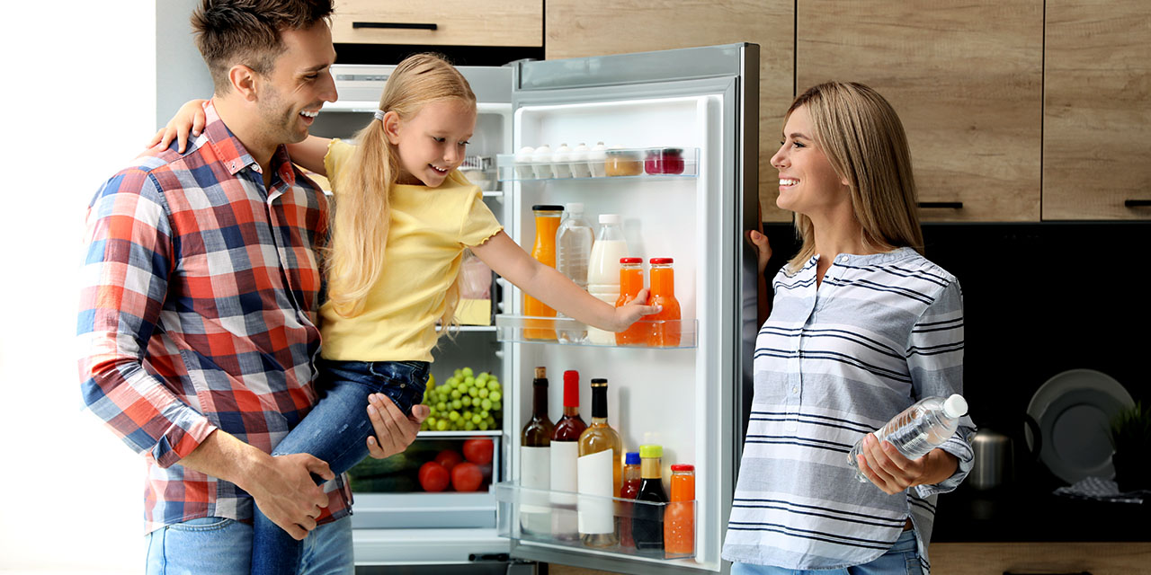 A family smiles in a modern kitchen as a dad holds his daughter while she reaches for a drink in an open refrigerator and mom stands nearby holding a water bottle
