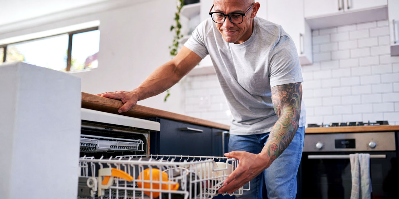 A man with glasses and a tattoo sleeve gently slides out a dishwasher rack in a bright, modern kitchen