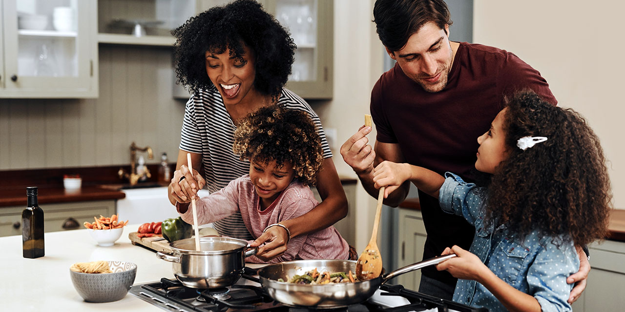 Family cooking together at the stove as kids help stir and taste in the kitchen