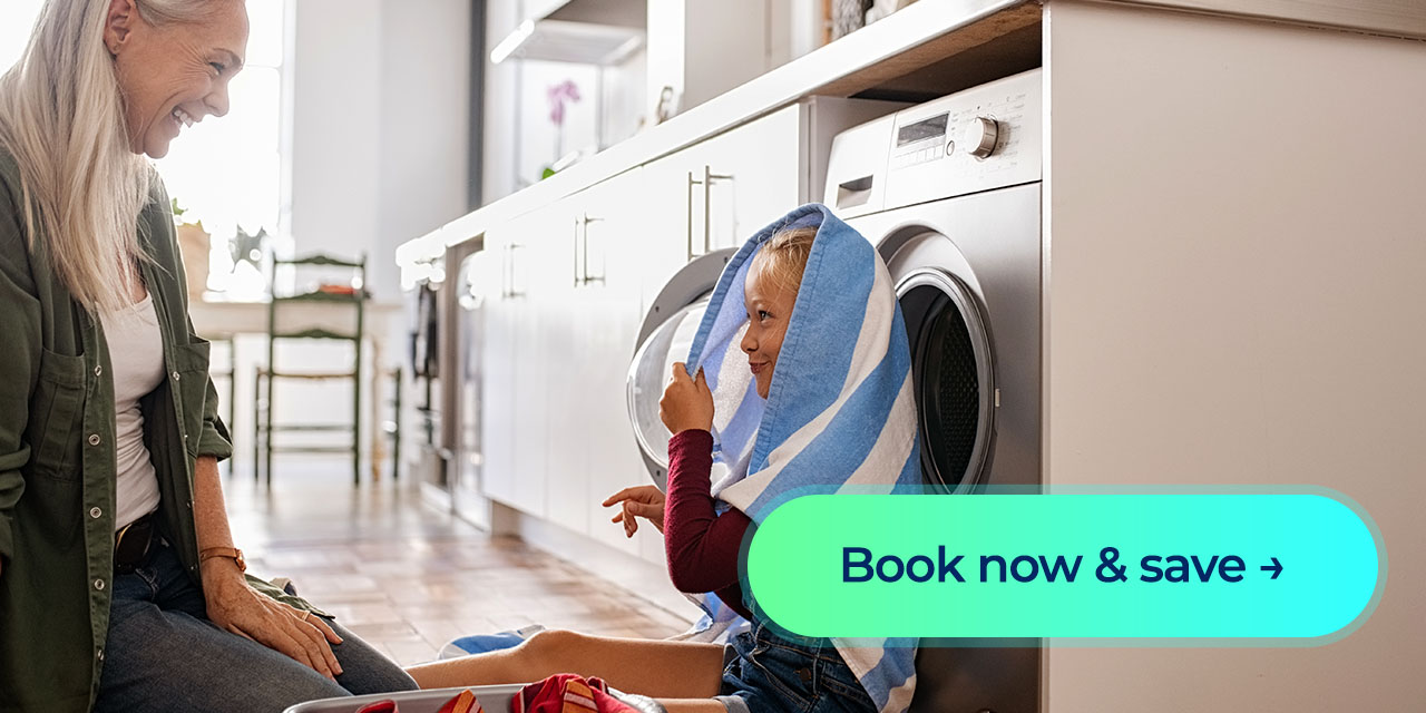 An older woman playing around near a washer with her granddaughter who has a towel over her head