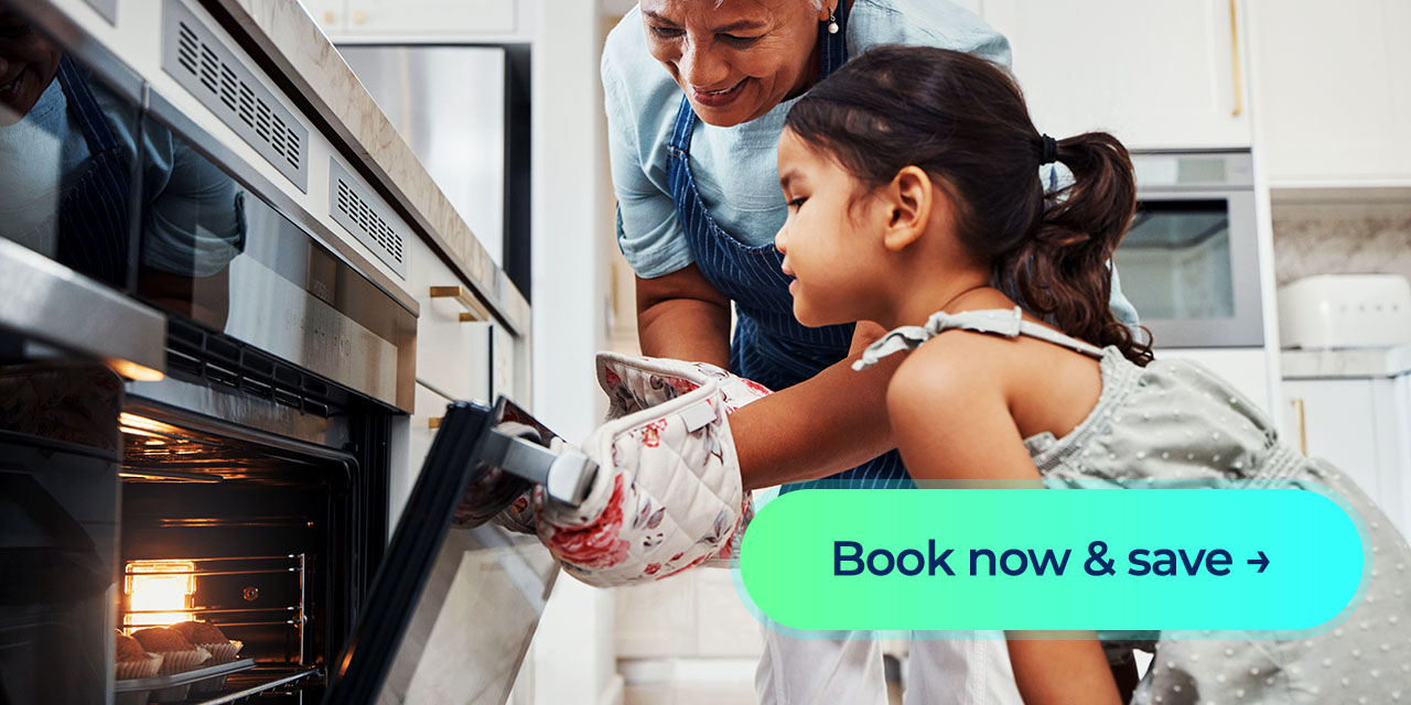 A grandmother pulls a tray of fresh muffins from the oven as her granddaughter watches with excitement beside a “Book now & save” button