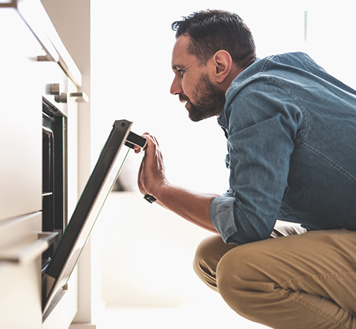 Man kneels in front of an open oven, checking inside to see how it is working