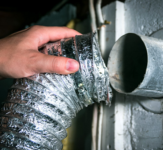 Hand holding a detached flexible dryer vent duct next to a wall exhaust pipe