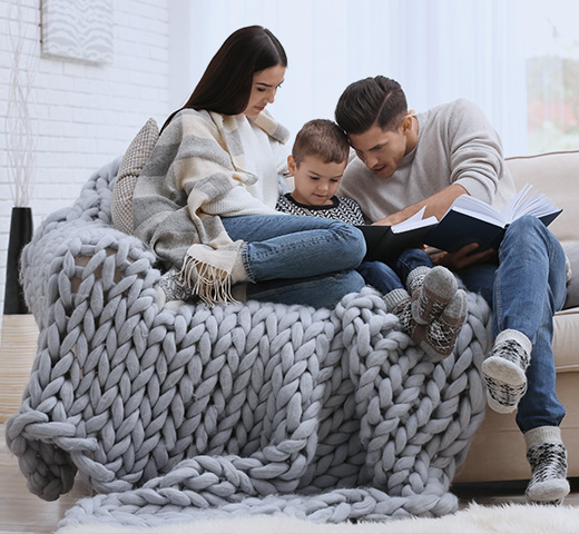 A family cuddled up on a beige couch with a grey blanket over them
