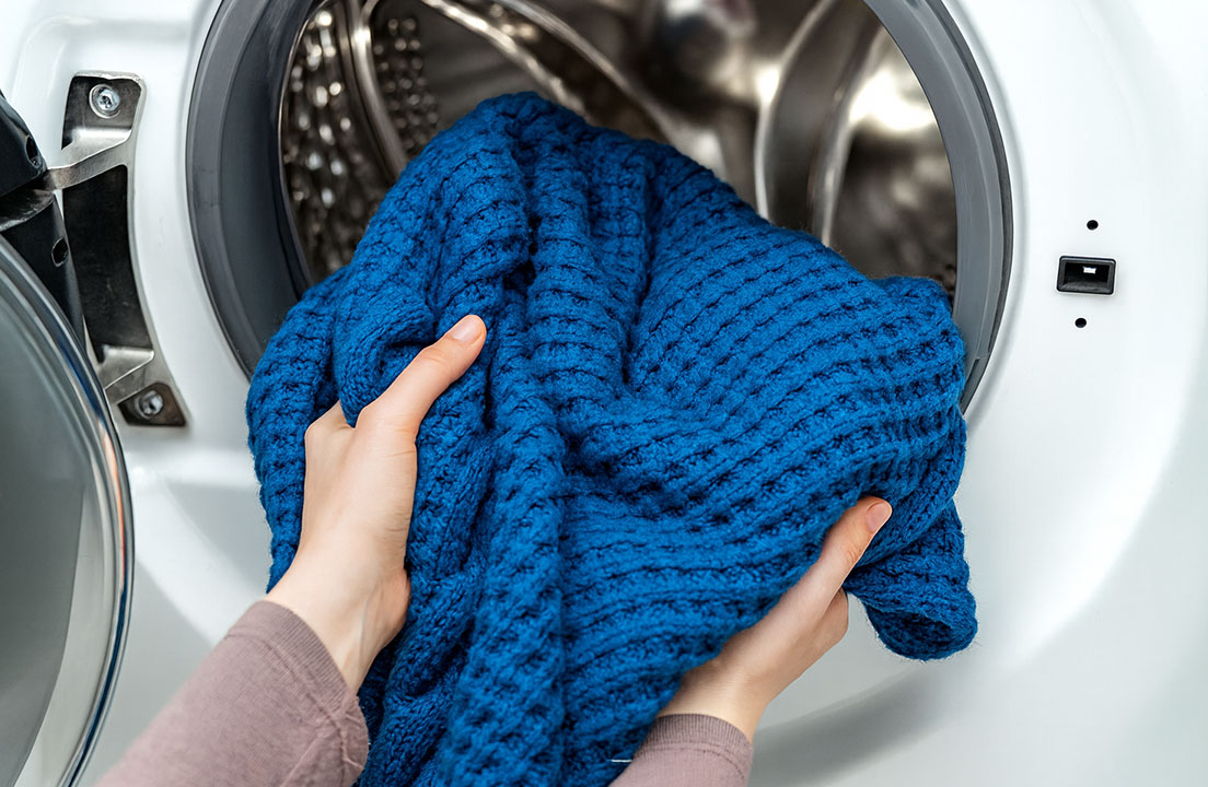 A person loads a thick blue winter blanket into a front-loading washing machine