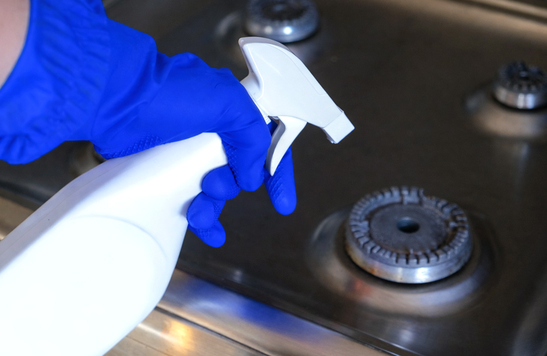 A hand in a blue cleaning glove holds a spray bottle over a stainless steel gas stovetop burner