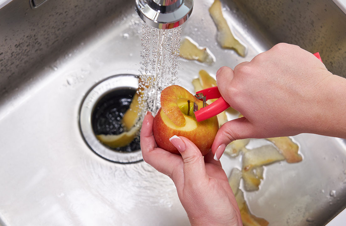 Hands peel an apple under running water above a kitchen sink and garbage disposal with apple peels scattered around the basin