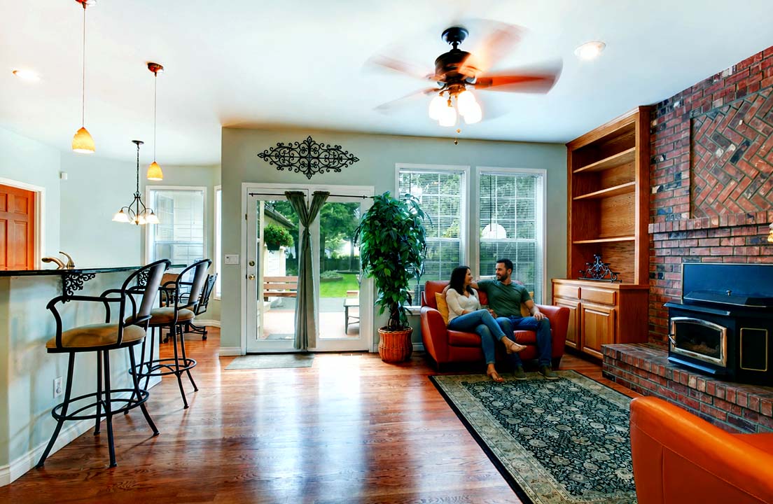 A couple relaxes indoors beneath a spinning ceiling fan in a sunlit living room