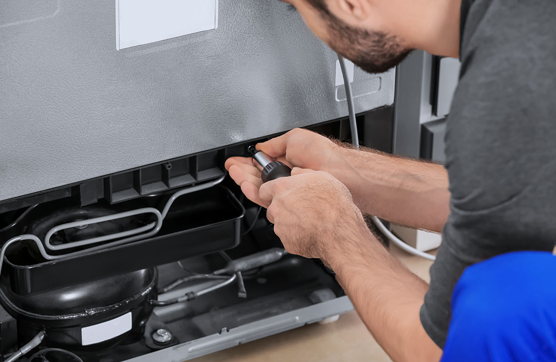 a technician kneeling in a kitchen behind a fridge unscrewing a panel