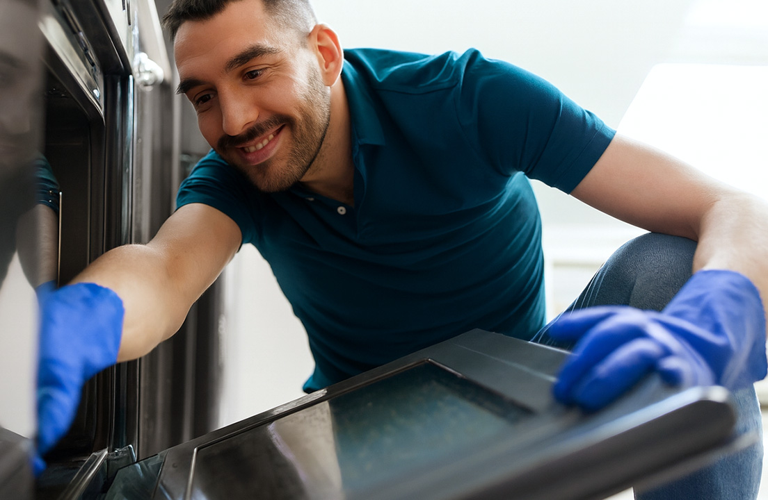 Smiling man wearing blue gloves kneels to carefully clean the inside of an open oven