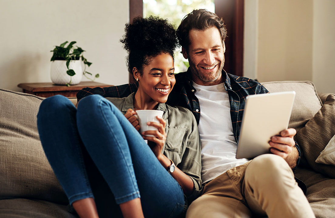 A couple sits on a couch smiling while looking at a tablet and holding a warm drink