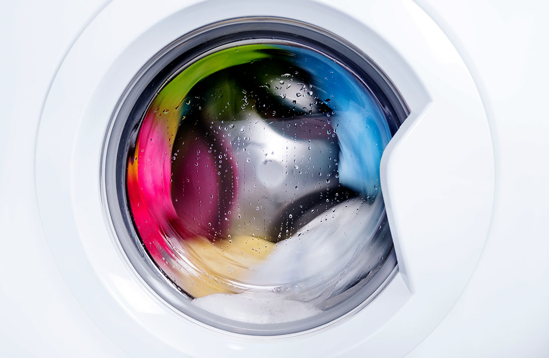 Colorful clothes tumble behind the glass door of a front load washer with water droplets