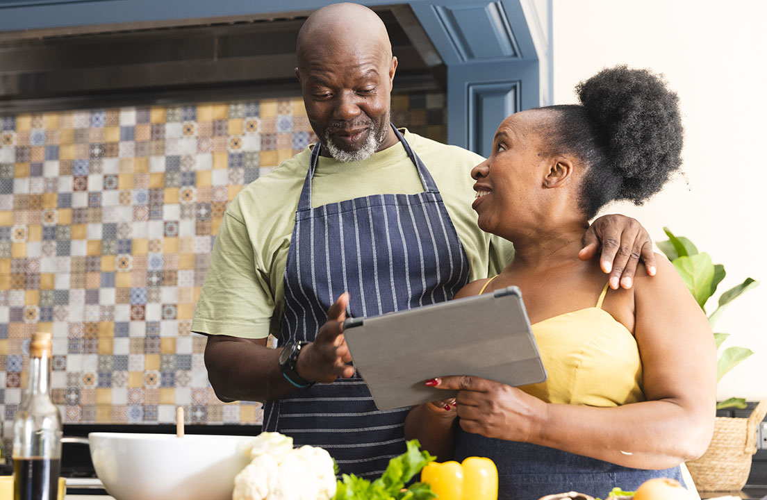 A couple in aprons stands in a kitchen looking at a tablet together with fresh vegetables and cooking ingredients on the counter