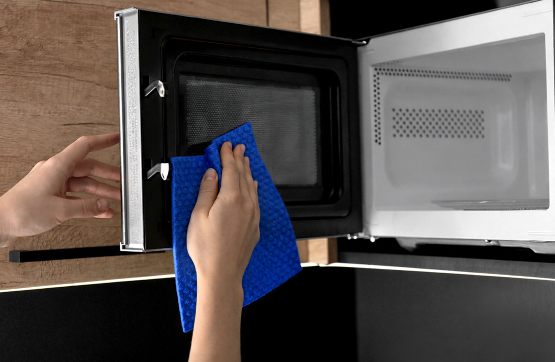 Person wiping the inside of an open microwave door with a blue cleaning cloth
