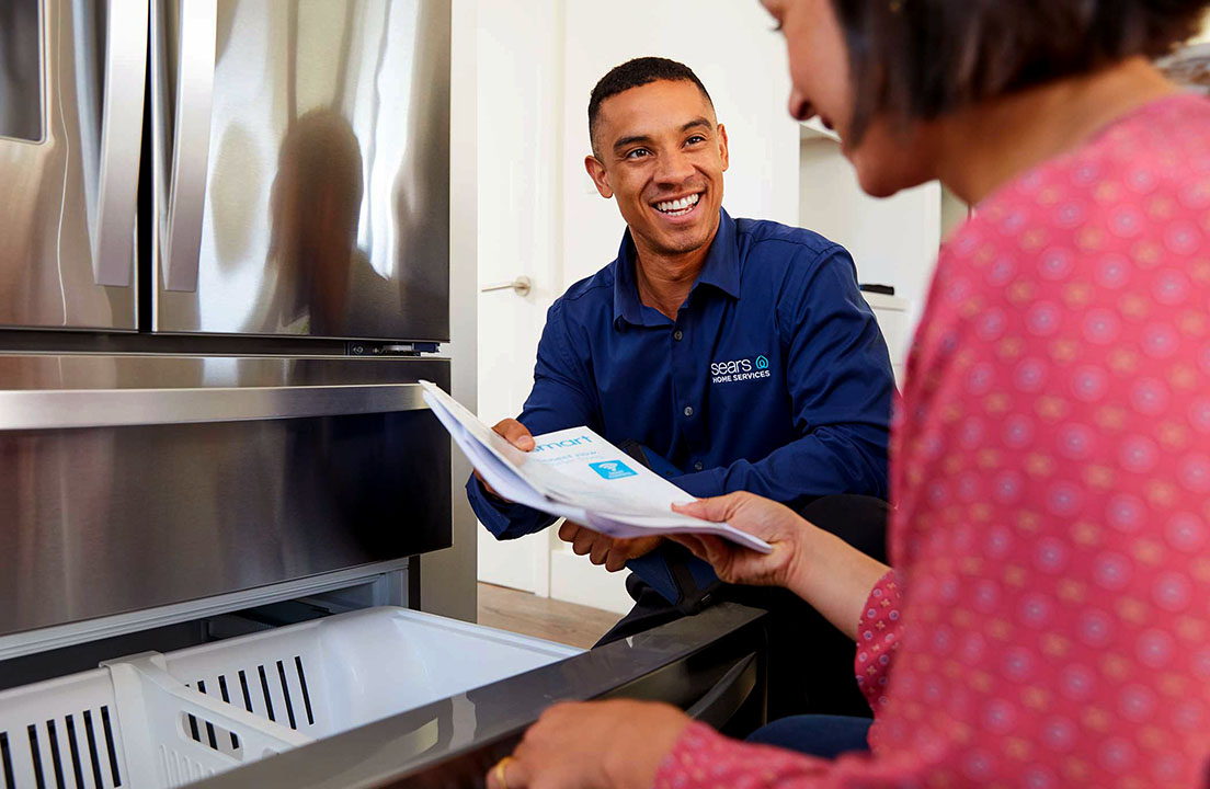 A Sears Home Services technician reviews home warranty paperwork with a homeowner near a refrigerator