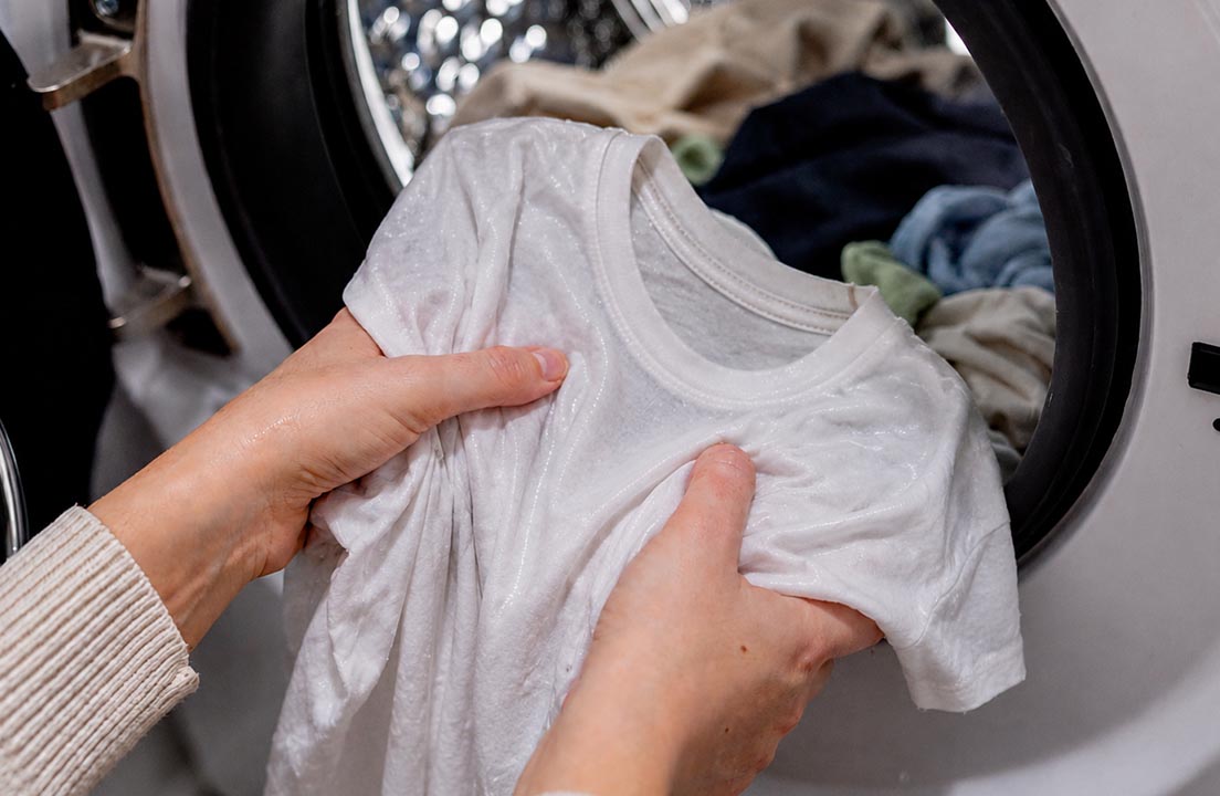 Hands hold a damp white shirt in front of an open dryer filled with clothes