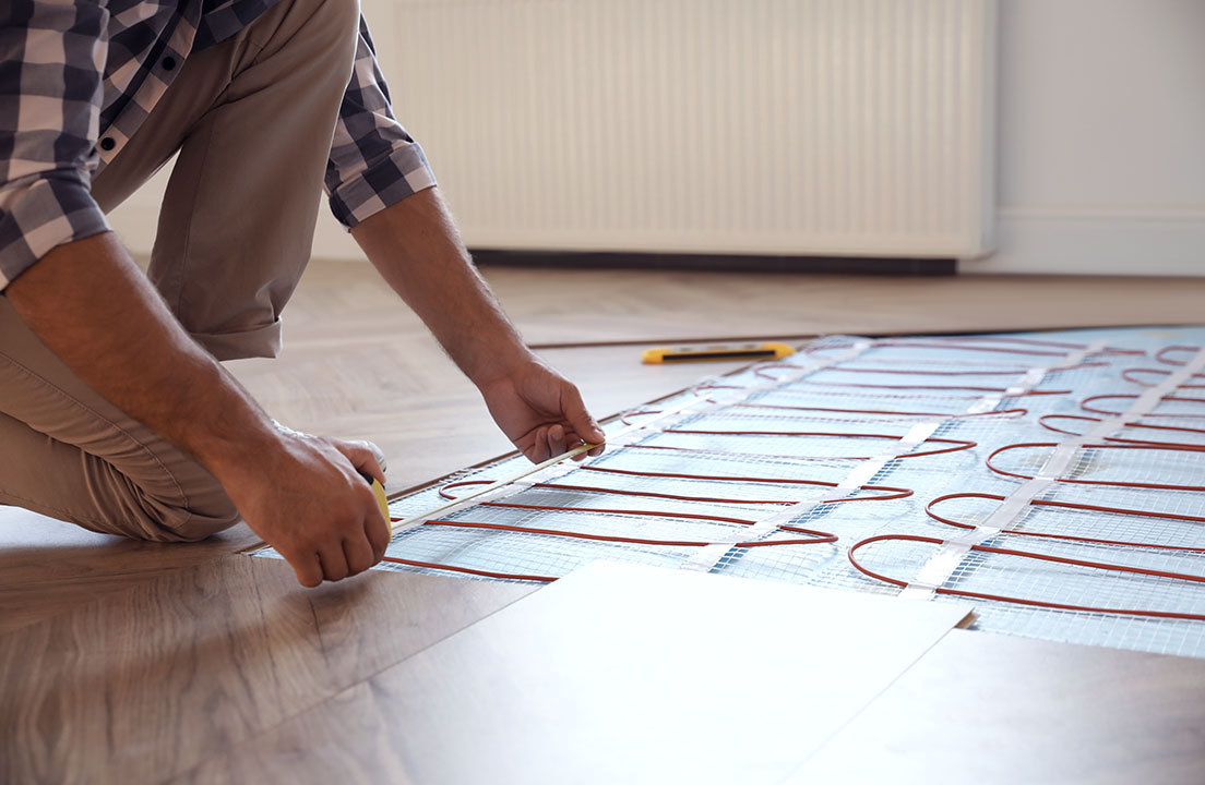 Person installing radiant floor heating coils beneath a wood floor, carefully measuring the layout for even home comfort