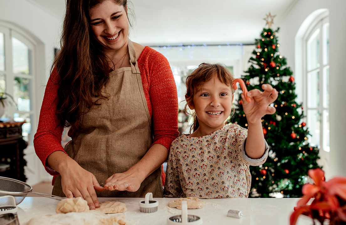 Mother and child happily shape holiday cookies at the kitchen counter while the child proudly holds a candy cane dough in front of a Christmas tree