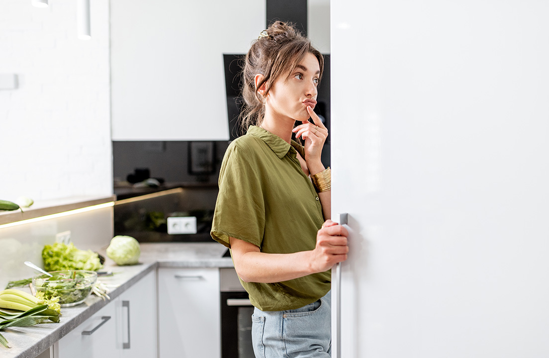 A woman stands at an open refrigerator, thinking while fresh vegetables sit on the kitchen counter