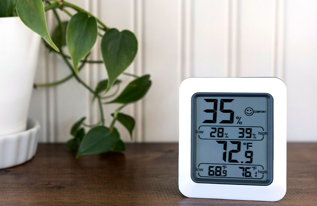 A digital humidity monitor on a table shows the indoor humidity level and temperature beside a plant