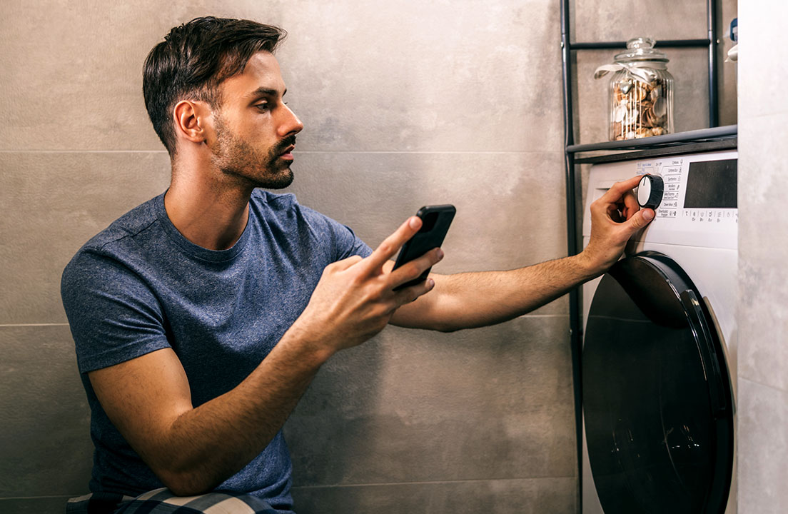 A man checks his phone while turning the dial on a front-loading washing machine