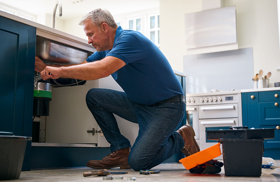 A man kneels on a kitchen floor repairing the plumbing beneath a sink with tools and a bucket nearby