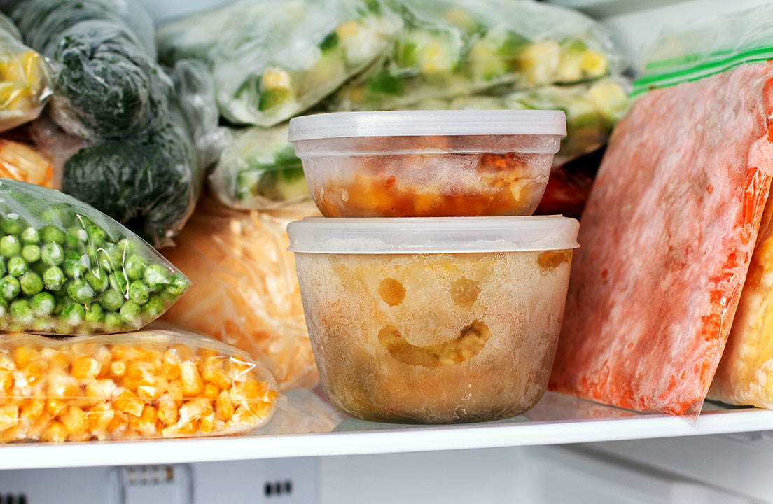 Frozen vegetables and containers fill a freezer, including a frosty food tub with a smiley face on the front