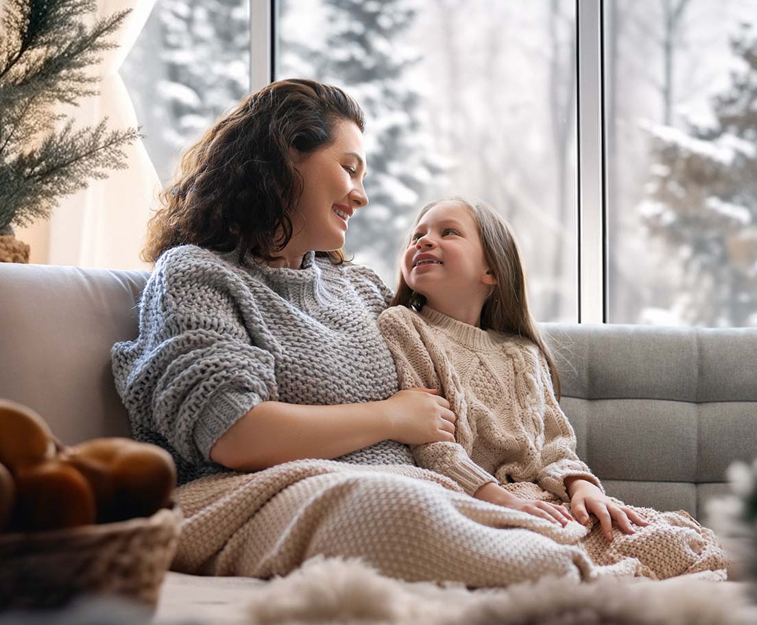Mother and daughter relax on a cozy couch in a warm living room while snow falls outside large windows