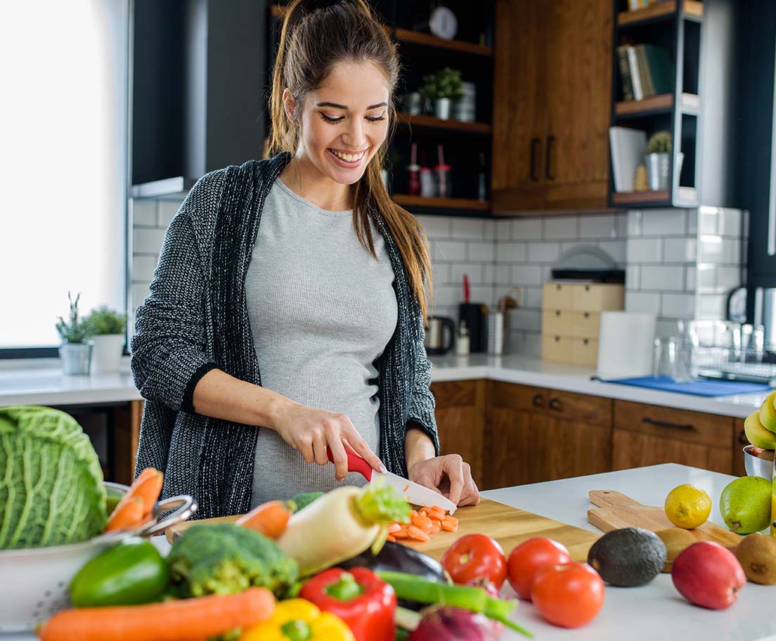 A smiling woman chops carrots on a cutting board in a kitchen surrounded by fresh produce