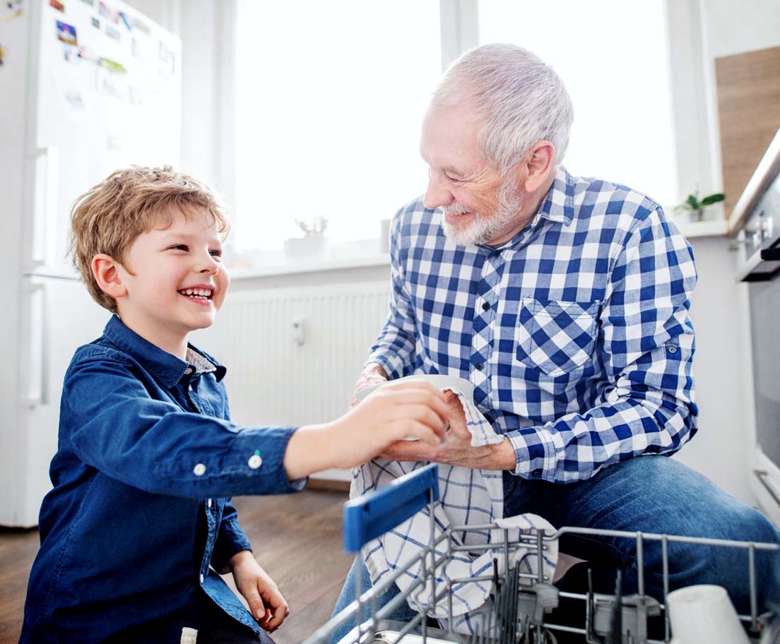 A smiling child hands a dish towel to an older man as they unload a dishwasher together in a bright kitchen