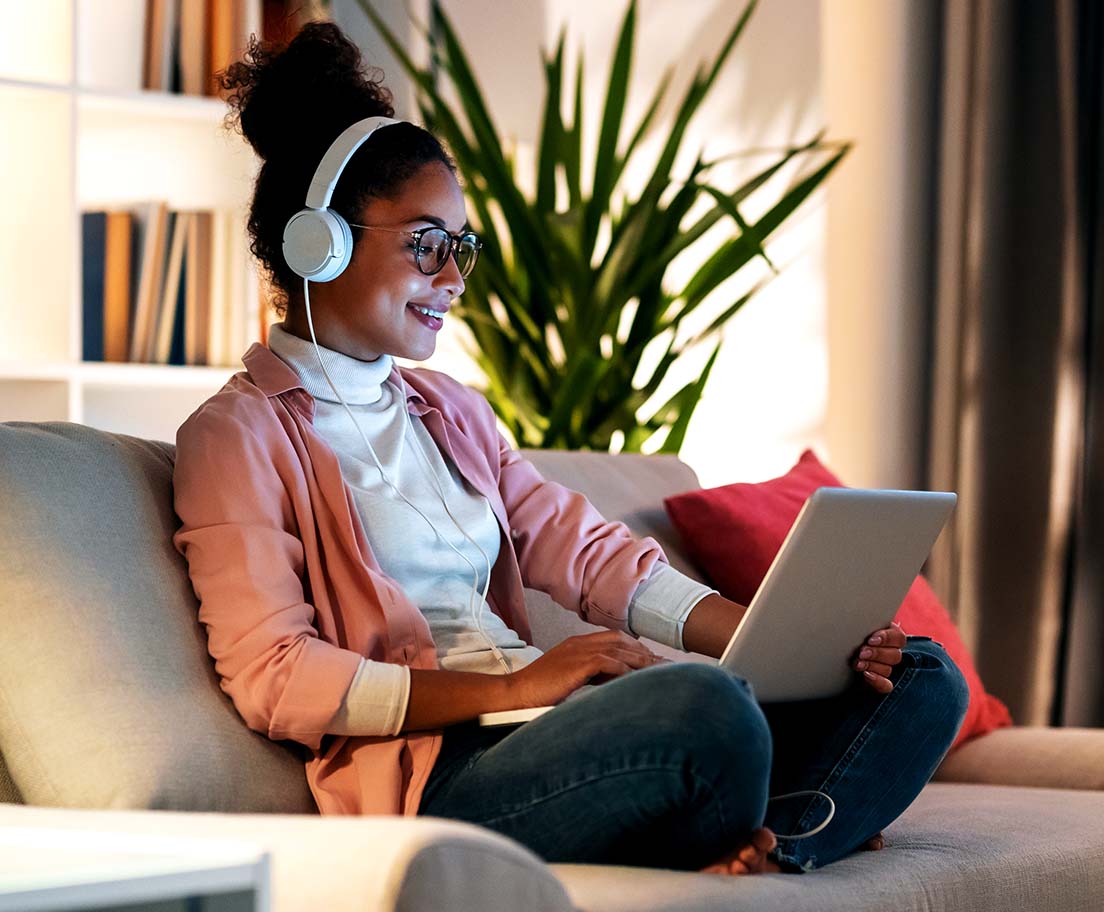 A woman in glasses and headphones sits on a couch using a laptop at home