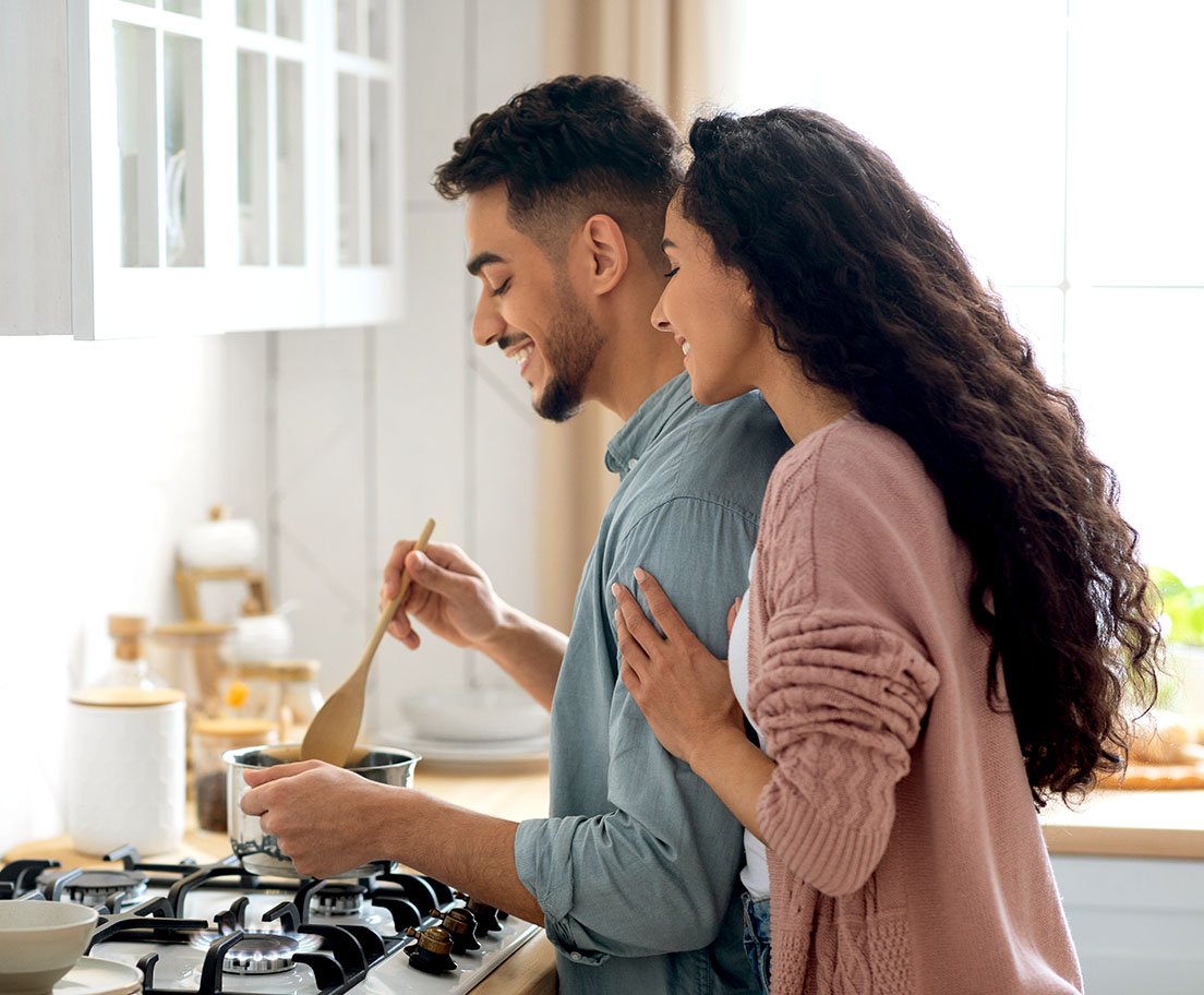A smiling couple stands at a gas stove in a bright kitchen while cooking together.