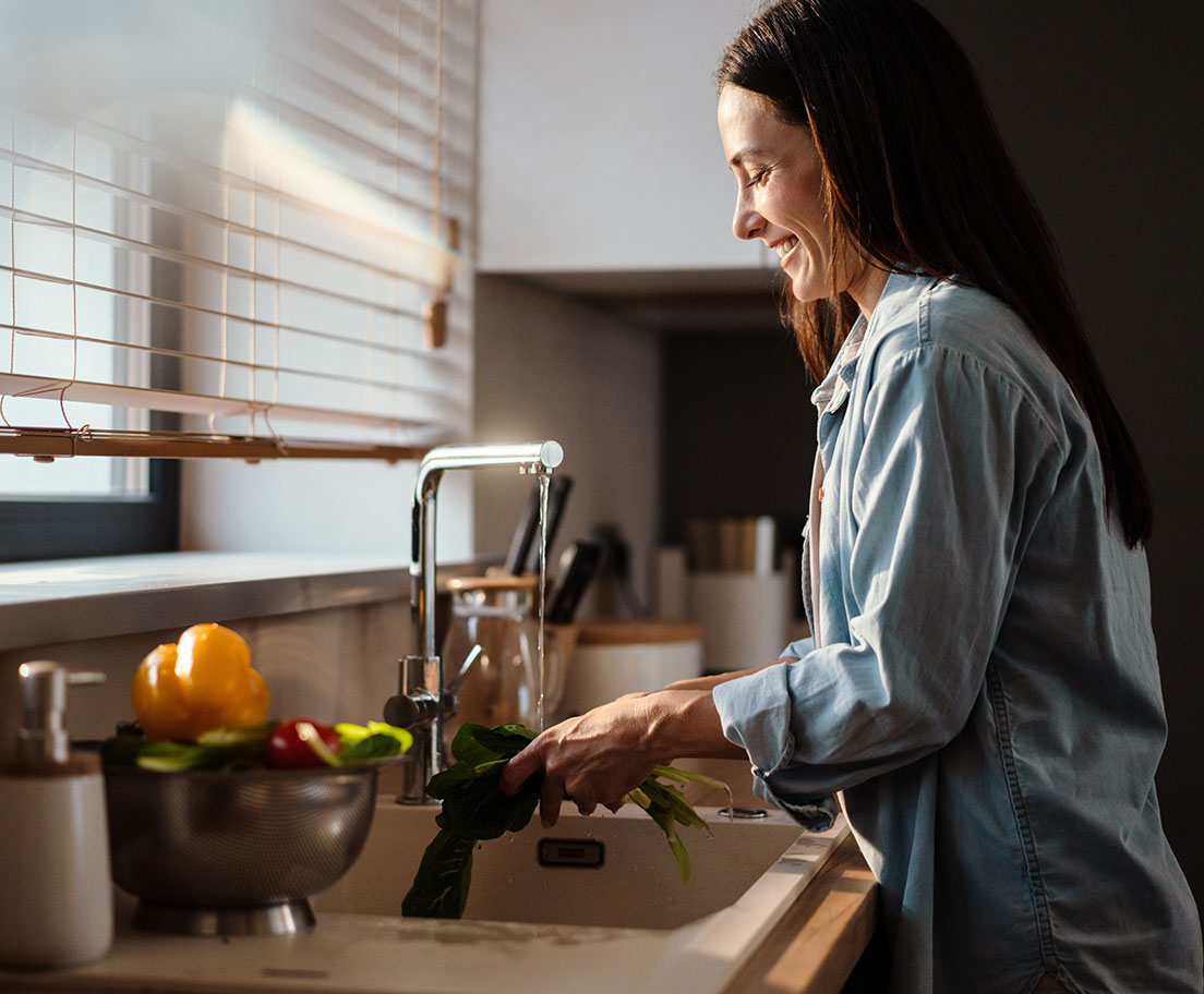 A smiling woman rinses leafy greens at a kitchen sink beside a sunlit window with fresh vegetables on the counter