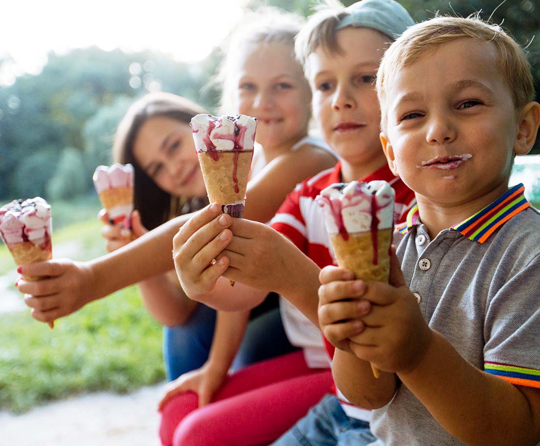 Four children sit together outdoors holding melting ice cream cones and smiling in the summer light
