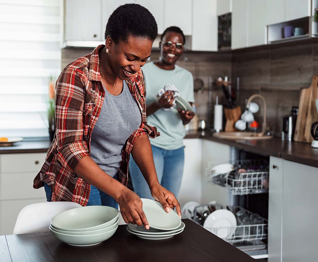 Two people unload clean dishes from an open dishwasher in a bright kitchen