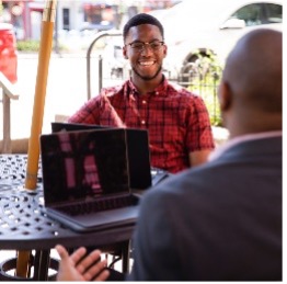Image of a mentor and a mentee talking outside at a table with a laptop between them